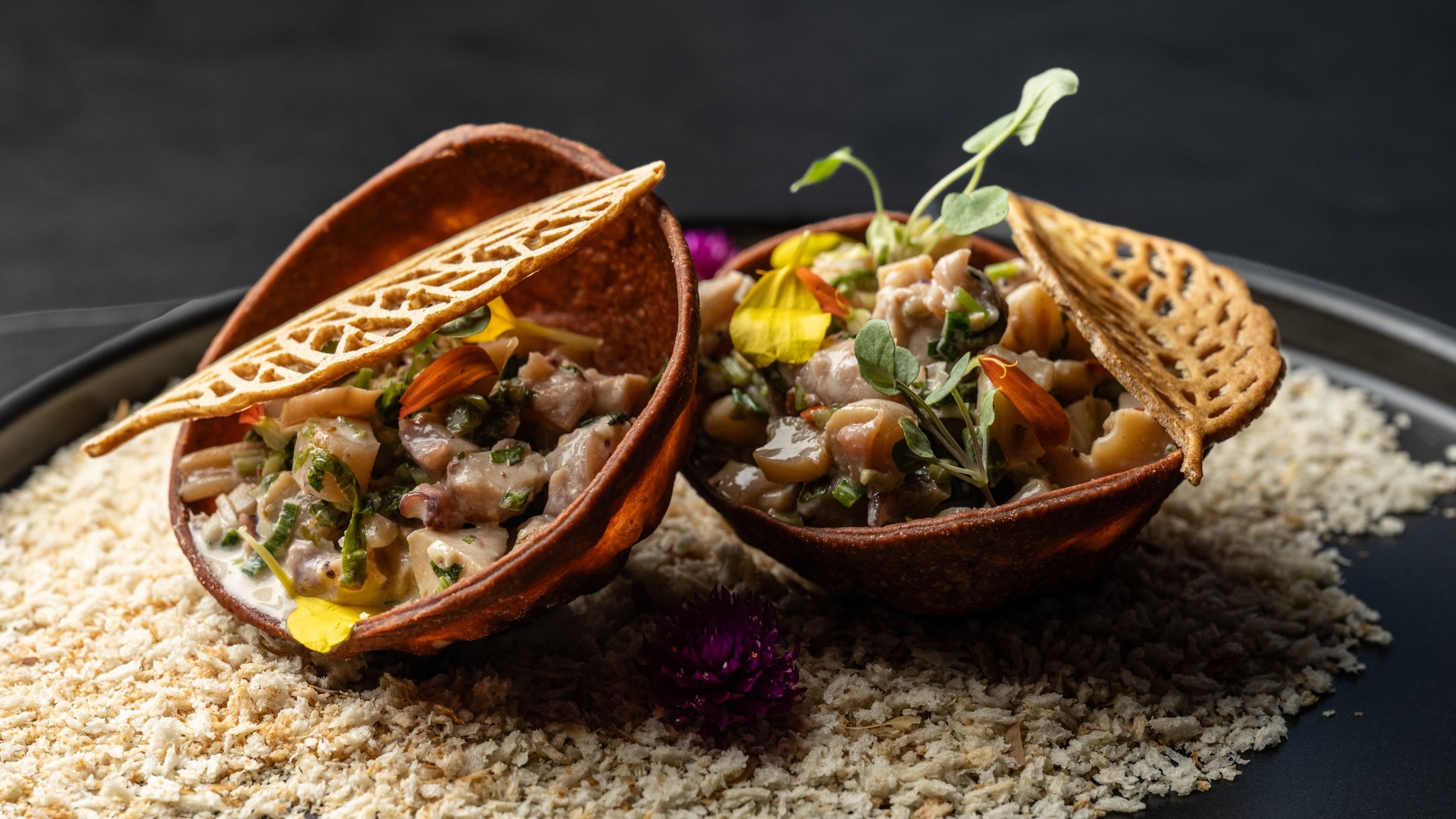 Dos tostadas de ceviche con flores comestibles y brotes, sobre un lecho de pan rallado en plato negro