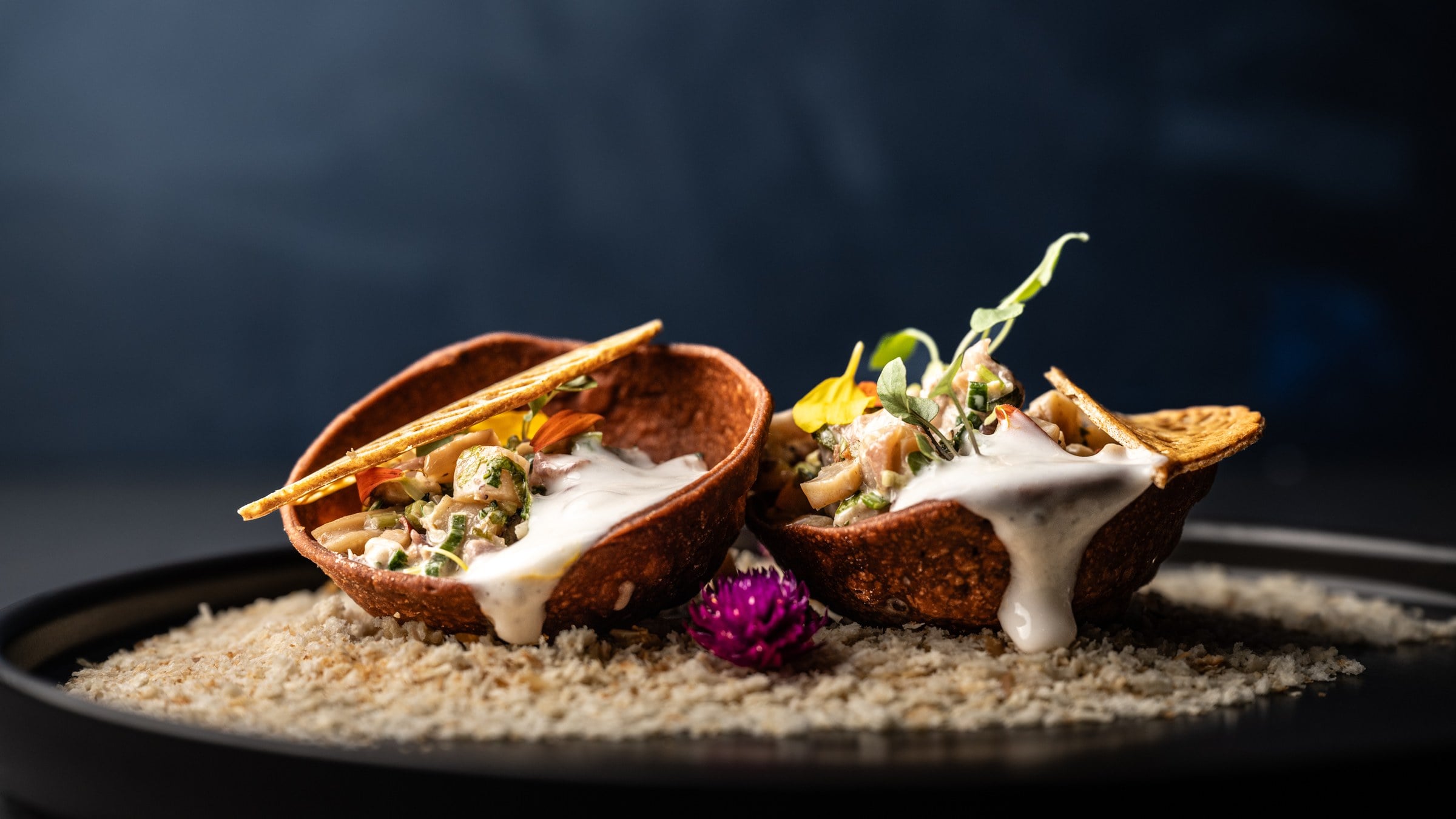 Ceviche servido en dos conchas crujientes con crema y una flor morada, sobre fondo azul oscuro